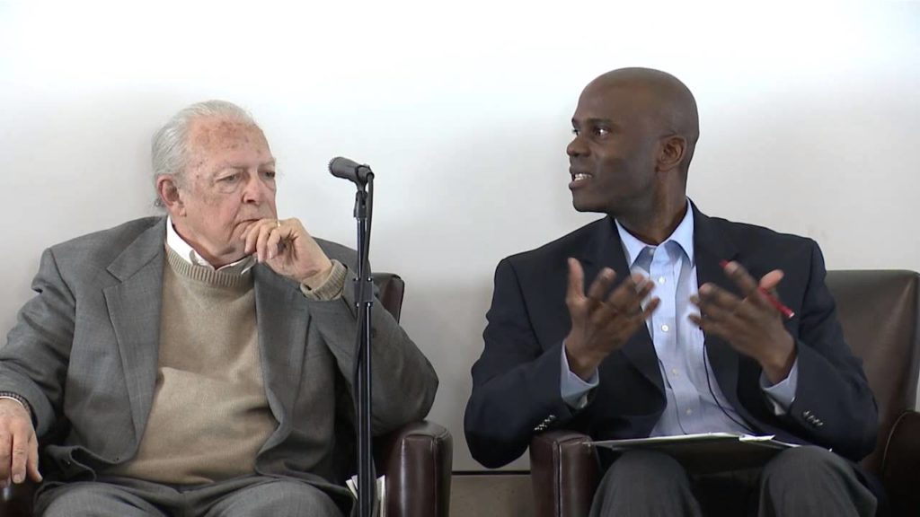 Two men sitting on a couch talking to each other.